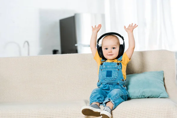 Очаровательный мальчик в наушниках с поднятыми вверх руками смотрит в камеру и слушает музыку дома — стоковое фото