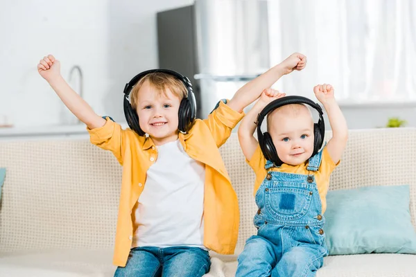 Adorable brothers in headphones looking at camera and cheering with hands in air at home — Stock Photo