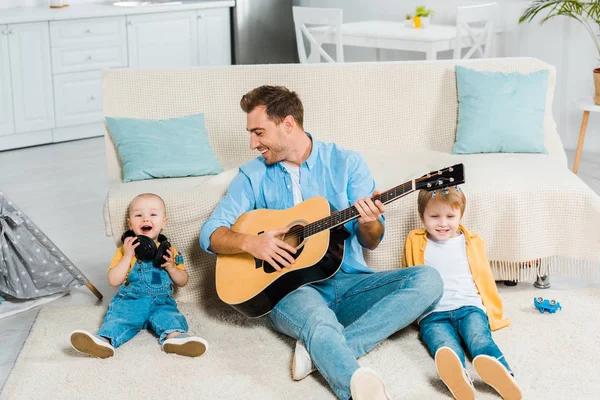 Улыбающийся отец играет на акустической гитаре для очаровательных счастливых дошкольников и маленьких сыновей дома — стоковое фото