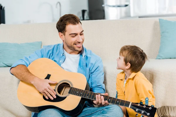 Bonito sorrindo pai tocando guitarra acústica para pré-escolar filho em casa — Fotografia de Stock