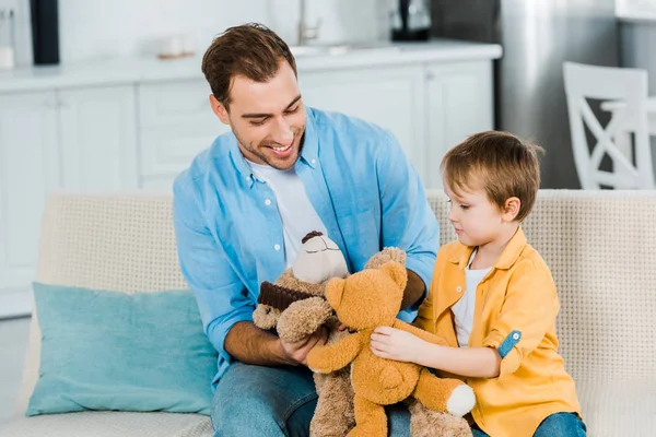 Pai e filho pré-escolar sentado no sofá e brincando com ursos de pelúcia em casa — Fotografia de Stock