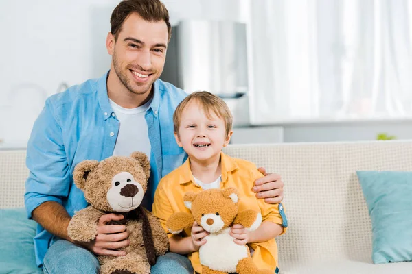 Padre e hijo preescolar sentado en el sofá y sosteniendo osos de peluche en casa - foto de stock