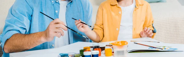 Vista recortada del padre y el hijo preescolar sosteniendo pinceles y dibujos en casa - foto de stock