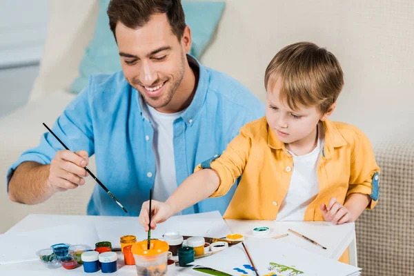 Smiling father and preschooler son holding paintbrushes and drawing at home — Stock Photo