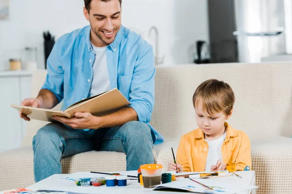 Smiling father reading book while cute preschooler son drawing at home — Stock Photo