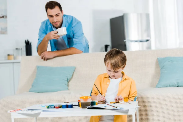 Preschooler son sitting on couch and drawing at home while father drinking coffee on background — Stock Photo