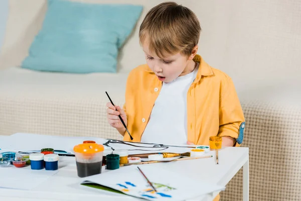 Surprised cute preschooler boy drawing with paintbrush at home — Stock Photo