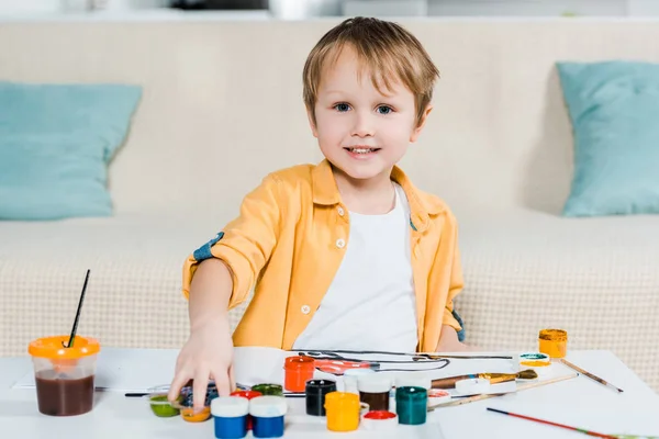 Entzückend lächelnder Vorschuljunge schaut in die Kamera, während er zu Hause zeichnet — Stockfoto