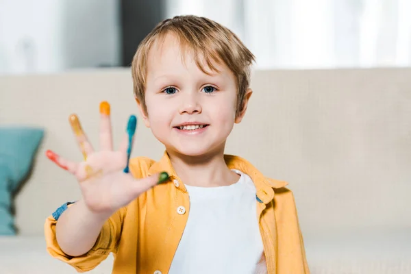 Adorable niño sonriente preescolar con pintura colorida en la mano mirando a la cámara en casa - foto de stock