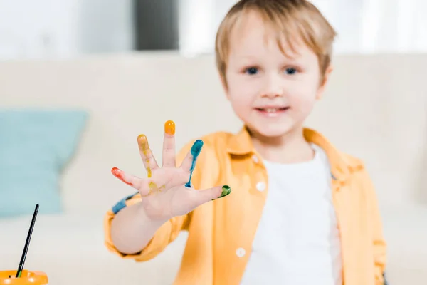 Mignon garçon préscolaire avec de la peinture colorée à portée de main en regardant la caméra à la maison — Photo de stock