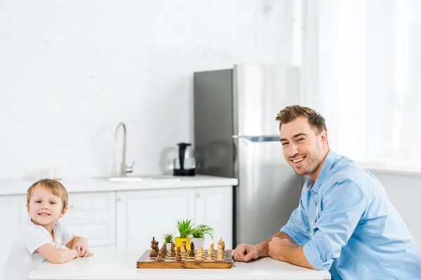 Heureux père et enfant d'âge préscolaire assis à table et regardant la caméra tout en jouant aux échecs dans la cuisine — Photo de stock