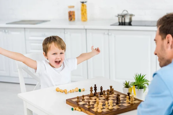 Bambino in età prescolare tifo con le mani in aria mentre gioca a scacchi con il padre a casa — Foto stock