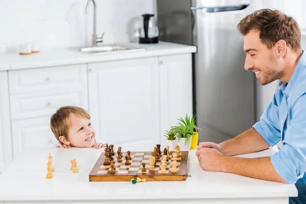 Улыбающийся отец и дошкольник играют в шахматы на кухне — стоковое фото