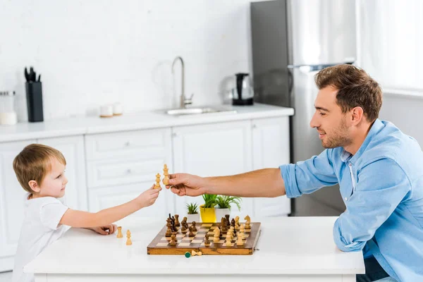 Vater und Sohn im Vorschulalter halten Schachfiguren, während sie am Tisch sitzen und zu Hause Schach spielen — Stockfoto