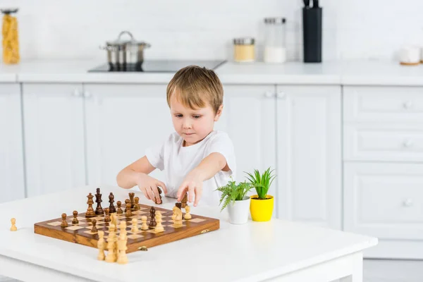 Adorable niño preescolar sentado en la mesa y jugando al ajedrez en casa - foto de stock