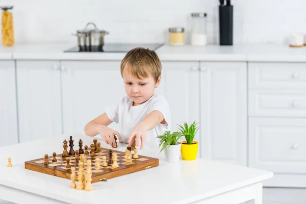 Очаровательный дошкольник сидит за столом и играет в шахматы дома — Stock Photo