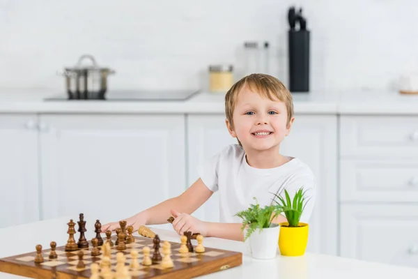 Entzückend lächelnder Vorschuljunge, der am Tisch sitzt, in die Kamera schaut und zu Hause Schach spielt — Stockfoto