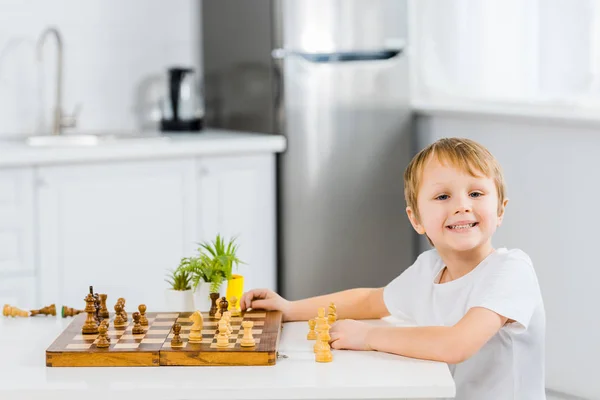 Adorable garçon préscolaire souriant assis à la table, regardant la caméra et jouant aux échecs à la maison — Photo de stock