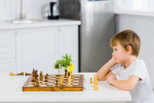 Очаровательный дошкольник сидит за столом и играет в шахматы дома — стоковое фото