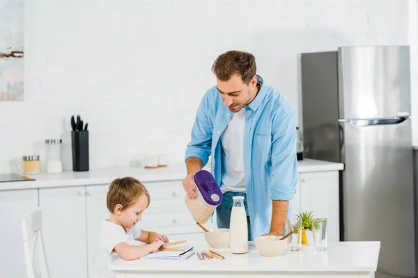 Handsome father pouring cereal in bowl while preschooler son drawing during breakfast in morning — Stock Photo