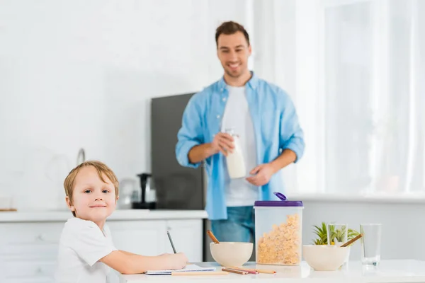 Bambino in età prescolare disegno e guardando la fotocamera durante la colazione con il padre sullo sfondo in cucina — Foto stock