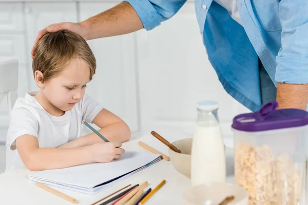 Père caressant préscolaire fils dessin pendant le petit déjeuner dans la cuisine — Photo de stock