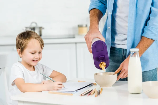 Батько наливає зерна в миску, коли посміхається дошкільнята син малює під час сніданку вранці — стокове фото