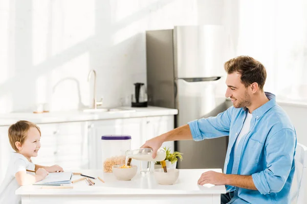 Pai derramando leite na tigela enquanto desenho pré-escolar durante o café da manhã na cozinha — Fotografia de Stock