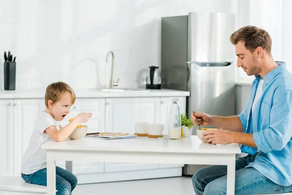 Padre e hijo preescolar comiendo cereales durante el desayuno en la cocina - foto de stock