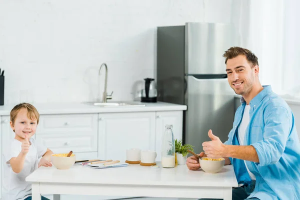 Glücklicher Vater und Vorschulsohn zeigen beim Frühstück in der Küche Daumen hoch und blicken in die Kamera — Stockfoto