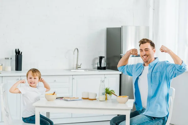 Sonriente hijo preescolar y padre mirando a la cámara, posando y mostrando bíceps durante el desayuno en la cocina - foto de stock