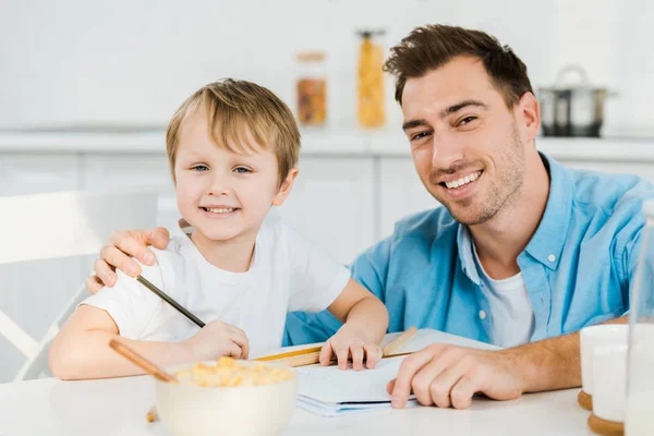 Padre e hijo preescolar mirando a la cámara y dibujo durante el desayuno en la cocina - foto de stock