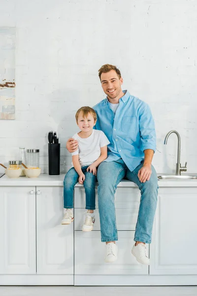 Souriant père et enfant d'âge préscolaire assis sur le comptoir et regardant la caméra pendant le petit déjeuner dans la cuisine — Photo de stock