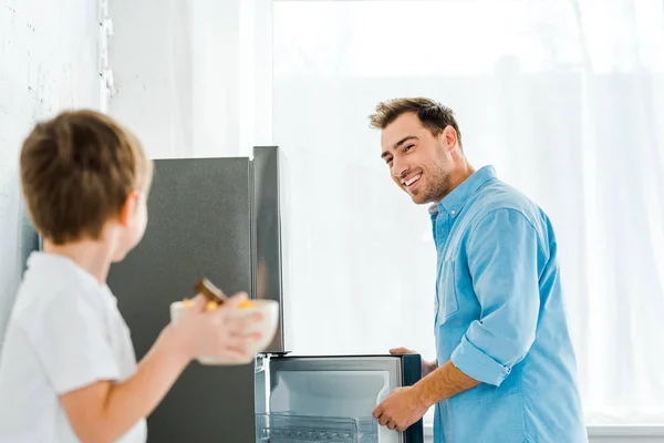 Messa a fuoco selettiva di padre sorridente in cucina con ragazzo che tiene ciotola di cereali in primo piano — Foto stock