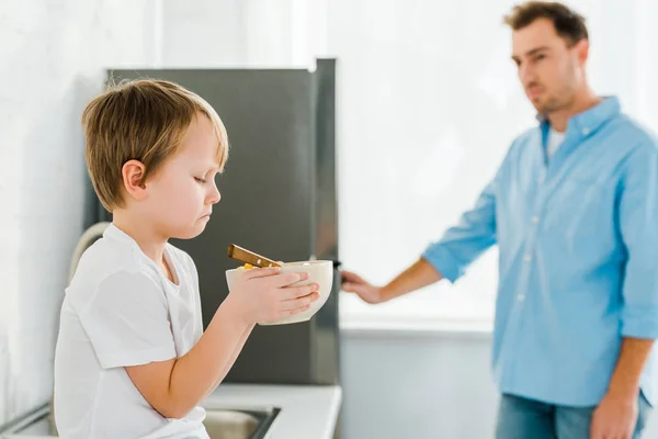 Sad boy holding bowl of cereal with disappointed father on background in kitchen — Stock Photo