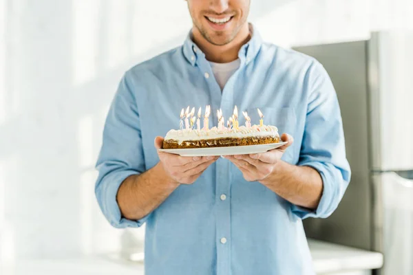 Vista recortada del hombre sonriente sosteniendo pastel de cumpleaños con velas encendidas en casa por la mañana - foto de stock