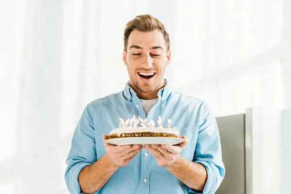 Збуджений красивий чоловік тримає торт на день народження з палаючими свічками вдома вранці — стокове фото
