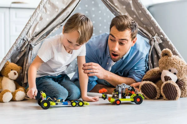 Sorprendido padre mirando preescolar hijo jugando con juguete coche bajo wigwam en casa - foto de stock