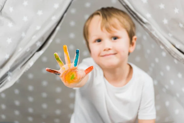 Cute preschooler boy with colorful paint on hand looking at camera at home — Stock Photo
