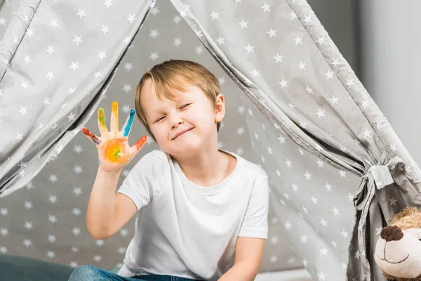 Adorable preschooler boy with colorful paint on hand sitting in wigwam at home — Stock Photo