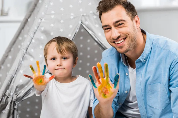 Heureux père et fils regardant caméra et montrant les mains peintes à la maison — Photo de stock