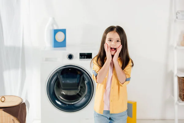 Frightened child in yellow shirt and jeans standing in laundry room — Stock Photo