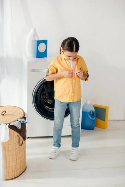 Niño de pie y desabrochando camisa amarilla cerca de lavadora y cesta en la sala de lavandería - foto de stock
