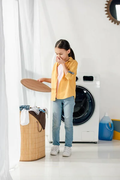 Child standing, holding basket cover wile feeling bad smell and covering nose with hand in laundry room — Stock Photo