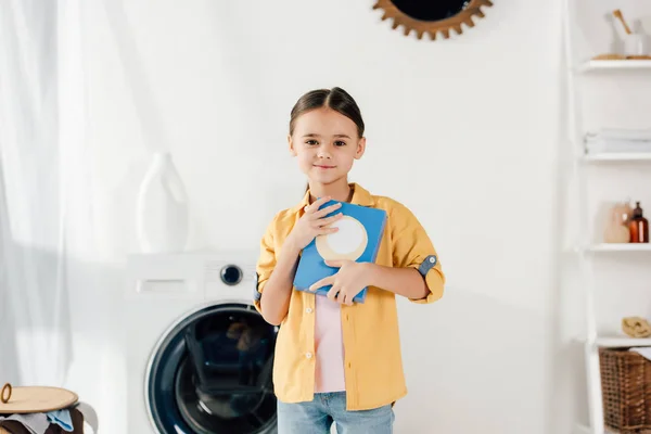 Kind steht neben Waschmaschine und hält Waschpulver in Waschküche — Stockfoto