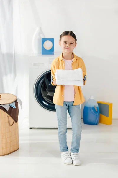 Kind steht neben Waschmaschine und hält weiße Handtücher in Waschküche — Stockfoto