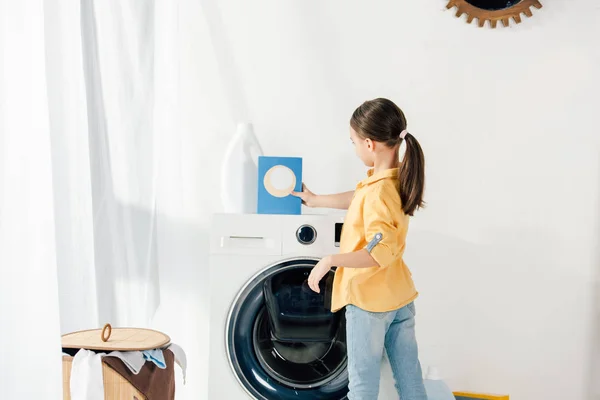 Side view of child in yellow shirt near washer taking washing powder in laundry room — Stock Photo