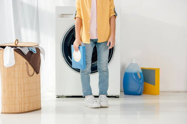 Cropped view of child in yellow shirt and jeans holding washing powder in laundry room — Stock Photo