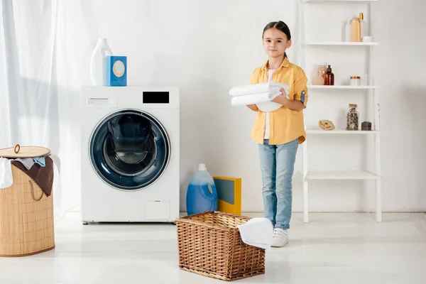 Child in yellow shirt near washer and ladder holding towels in laundry room — Stock Photo