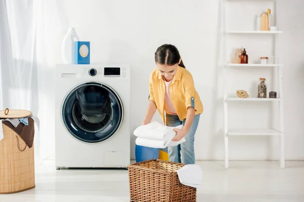 Kind in gelbem Hemd neben Waschmaschine und Leiter legt Handtücher in Korb in Waschküche — Stockfoto